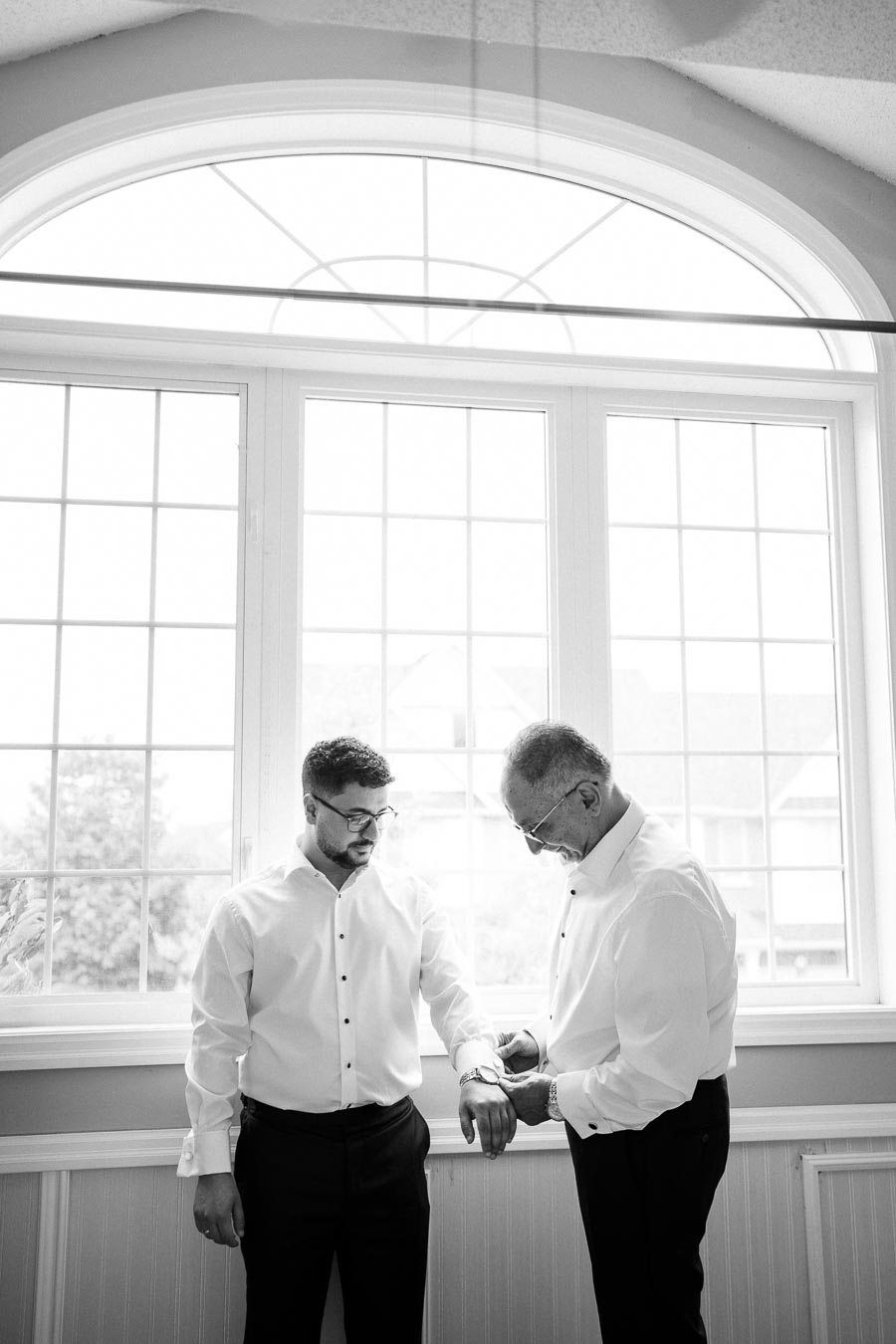 Two men in formal attire standing by a bright window, one assisting the other with a watch, creating a moment of preparation and bonding. Black and white photography.