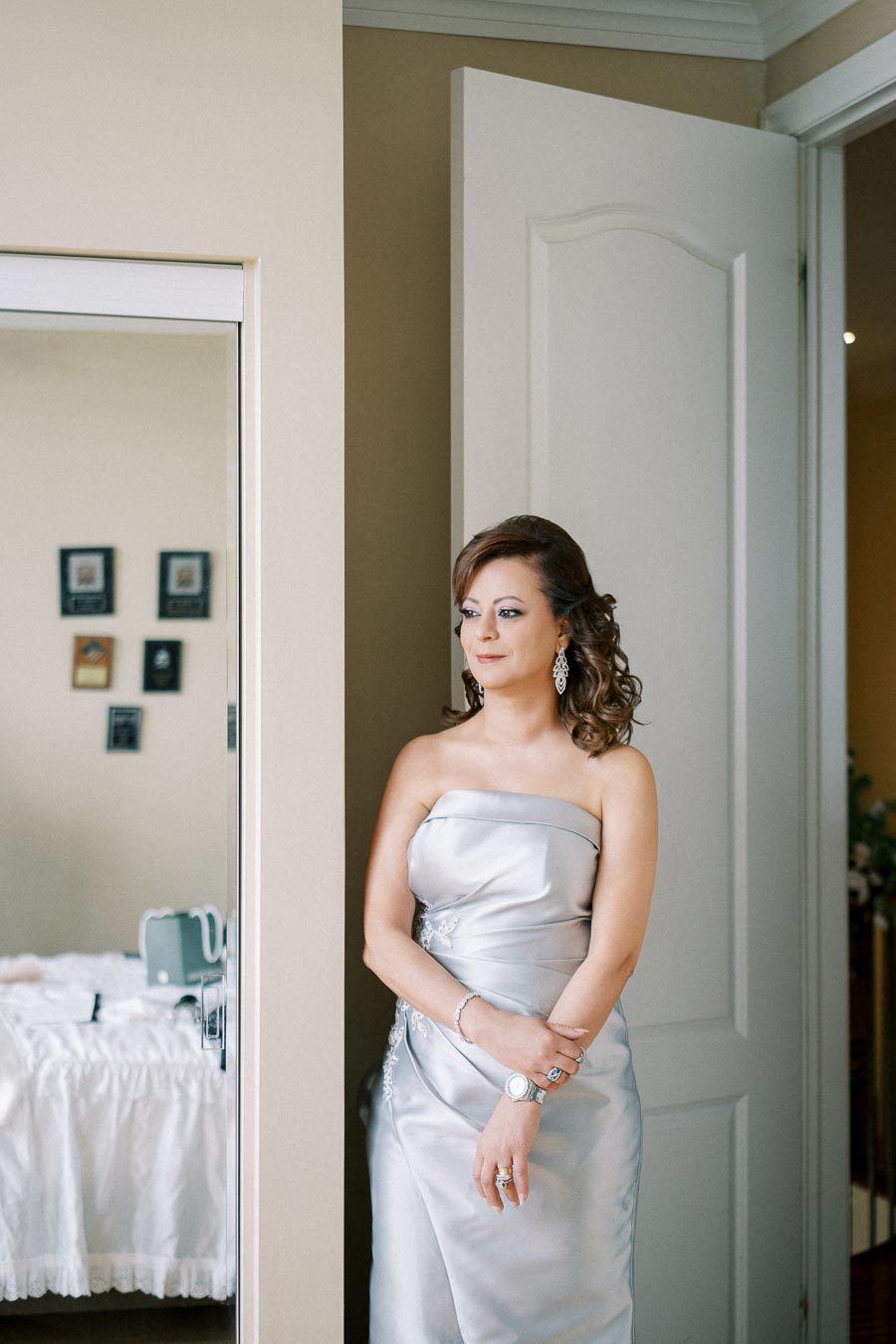 Elegant woman in a strapless silver dress standing beside a door, with a thoughtful expression and curly hairstyle, wearing silver jewelry and looking out into the room.