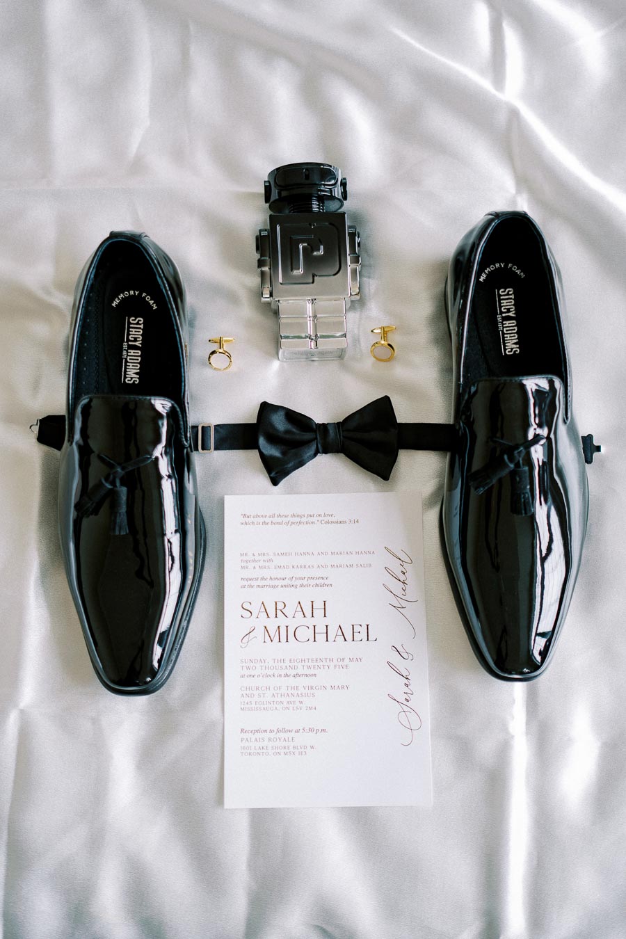 Elegant wedding accessories arrangement featuring glossy black dress shoes, a black bow tie, gold cufflinks, a sleek cologne bottle, and a wedding invitation with the names Sarah and Michael on a white satin background.
