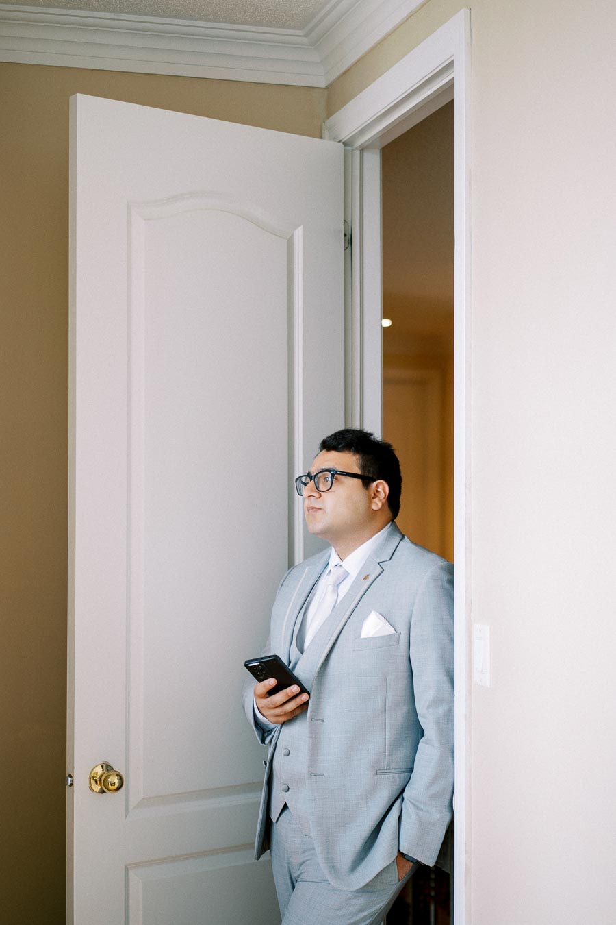 Man in light gray suit standing in a doorway, holding a smartphone and looking thoughtful.