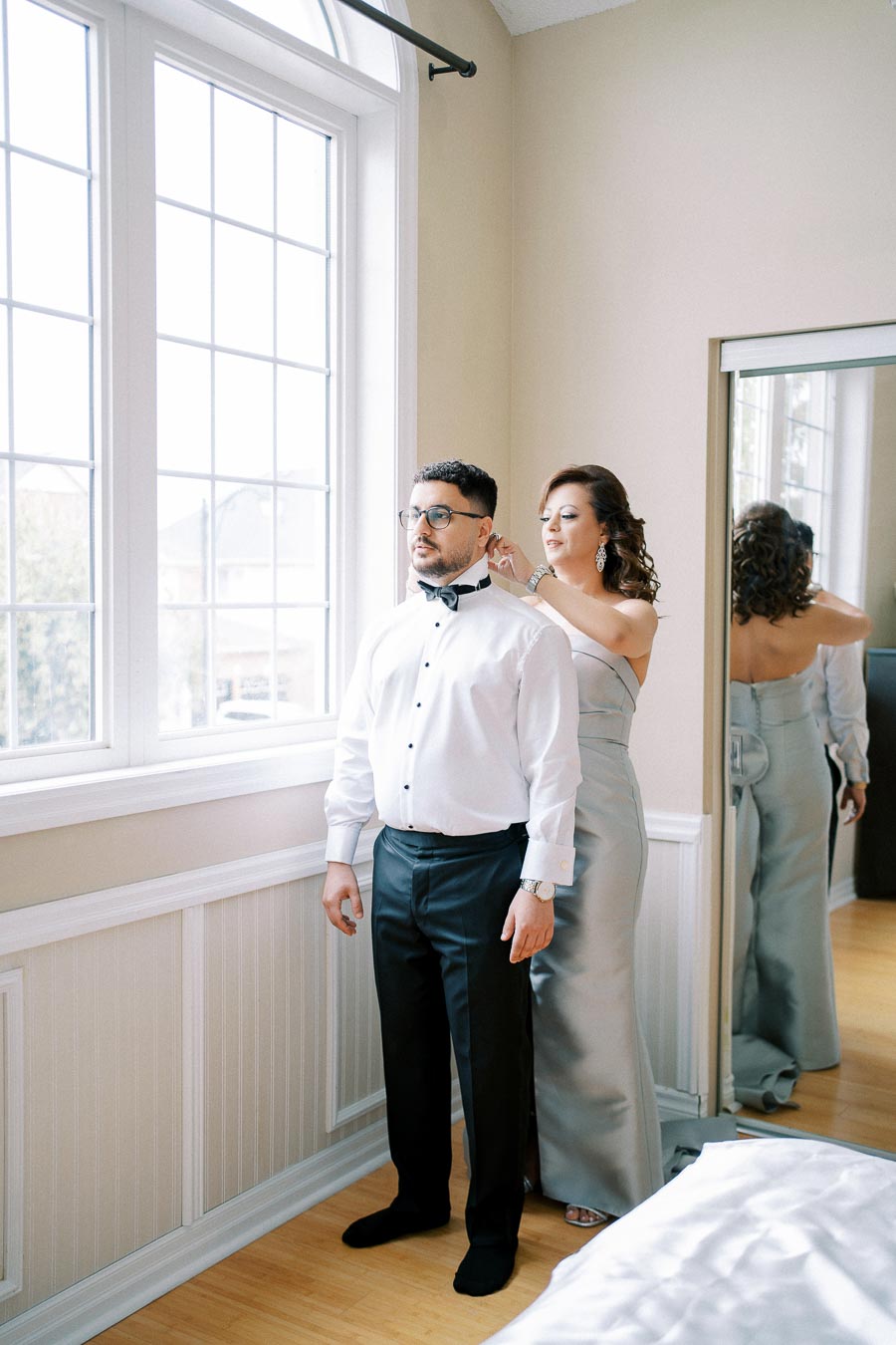 A woman in an elegant dress adjusts a bow tie for a man in formal attire by a large window, reflecting their preparation for a special event or wedding day.