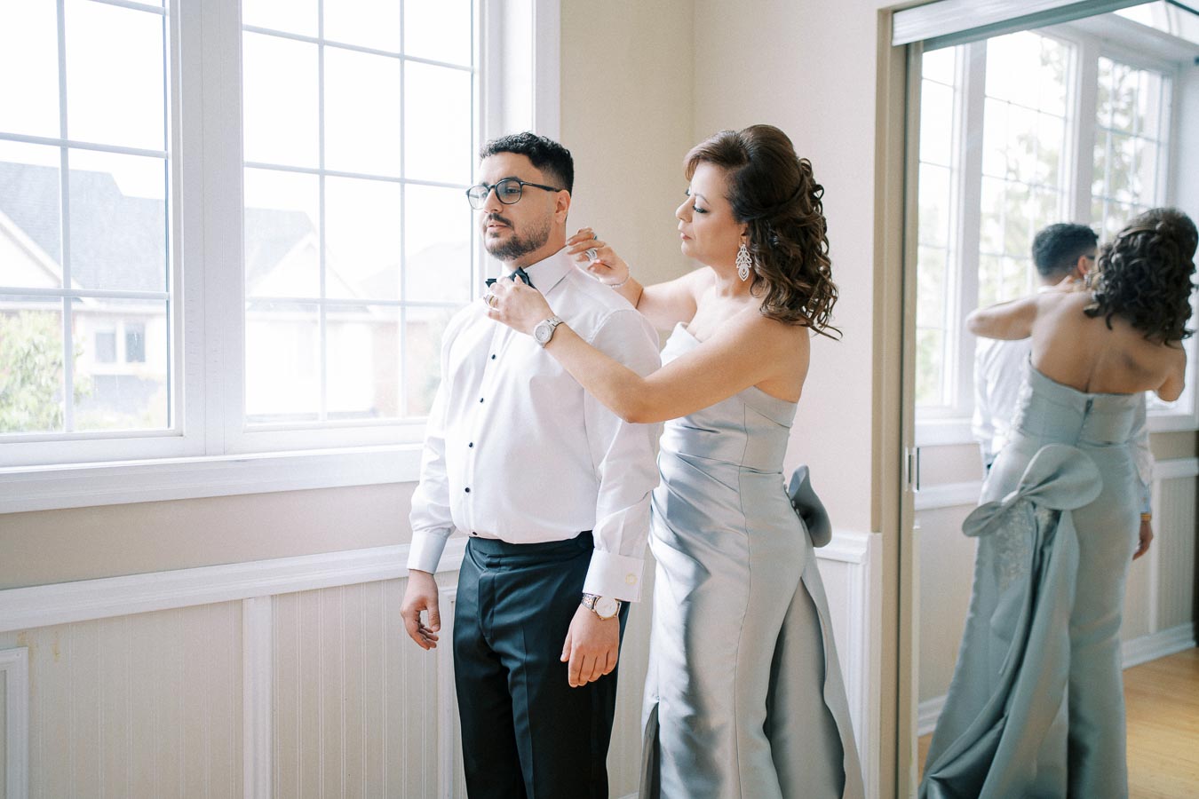 A woman in an elegant silver gown adjusts a man's bow tie in a bright room with large windows.