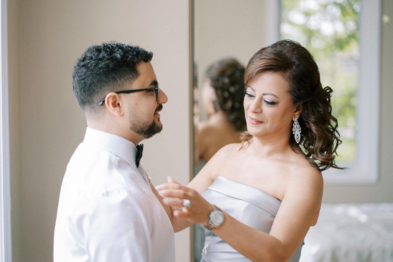 A woman in an elegant dress adjusts a man's bow tie while smiling, captured in a softly lit room with a window in the background.