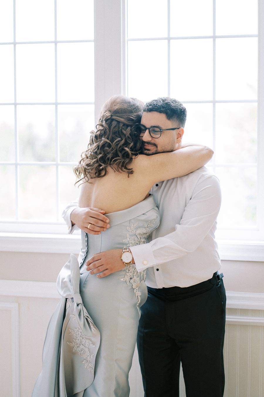 A couple embracing in front of a large window, with the woman wearing an elegant light blue dress with embroidery and the man in a white dress shirt with a watch, conveying love and happiness.