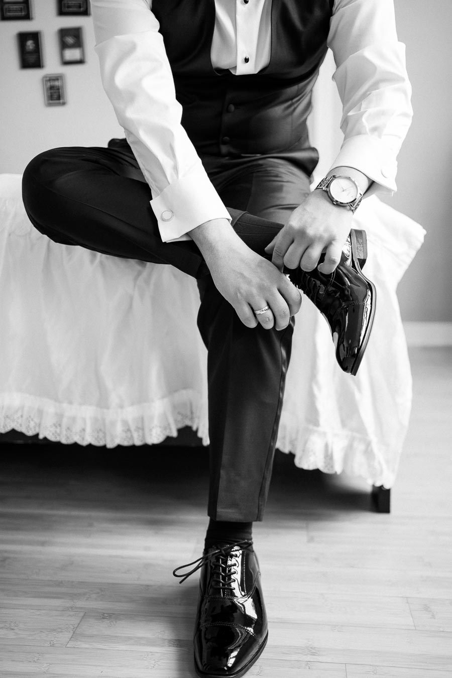 Black and white image of a groom dressing in formal attire, tying his sleek black leather shoes while sitting on a bed.