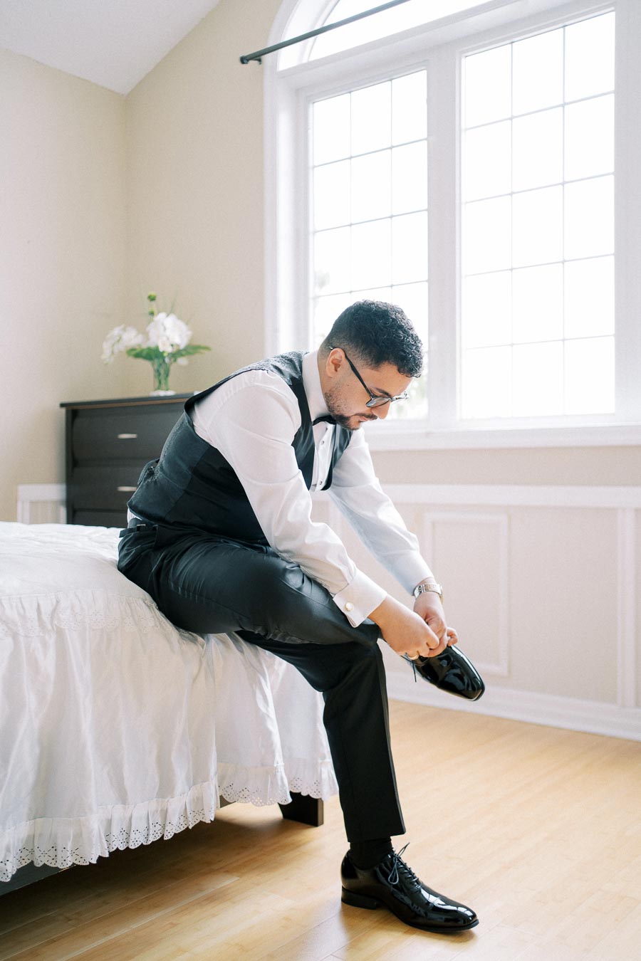 Man in formal attire sitting on a bed tying shoelaces in a bright room with large window and floral decor.