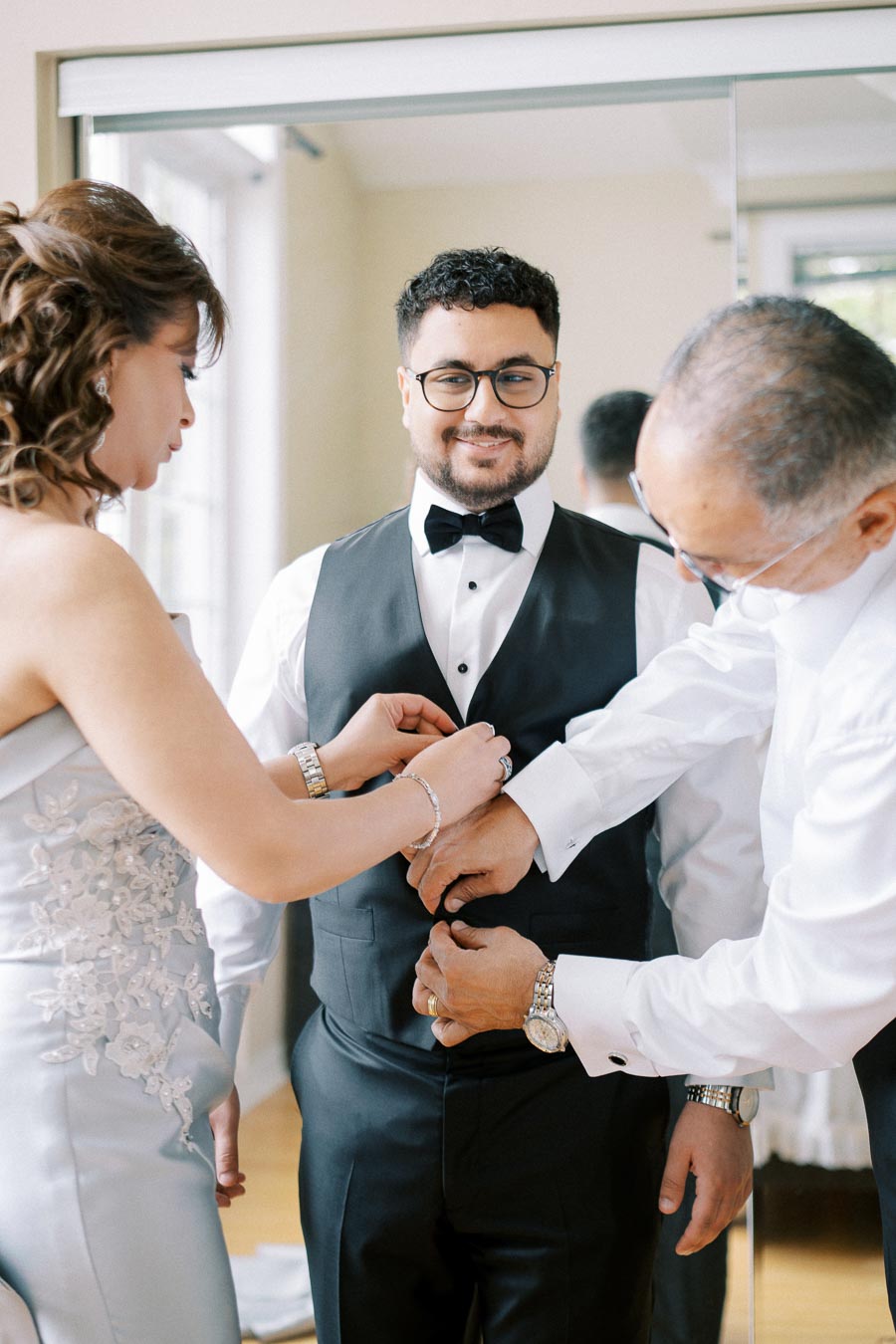 Groom smiling as he gets ready for wedding, assisted by family members adjusting his vest and bow tie in a bright room.