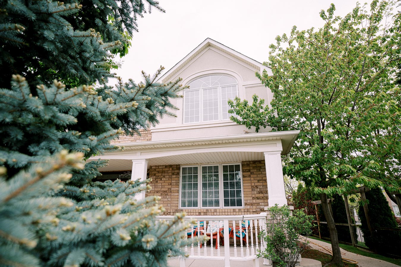 Alt text: Front view of a two-story house with a brick facade and large windows, surrounded by green foliage and a pine tree in the foreground, showcasing a welcoming porch area.