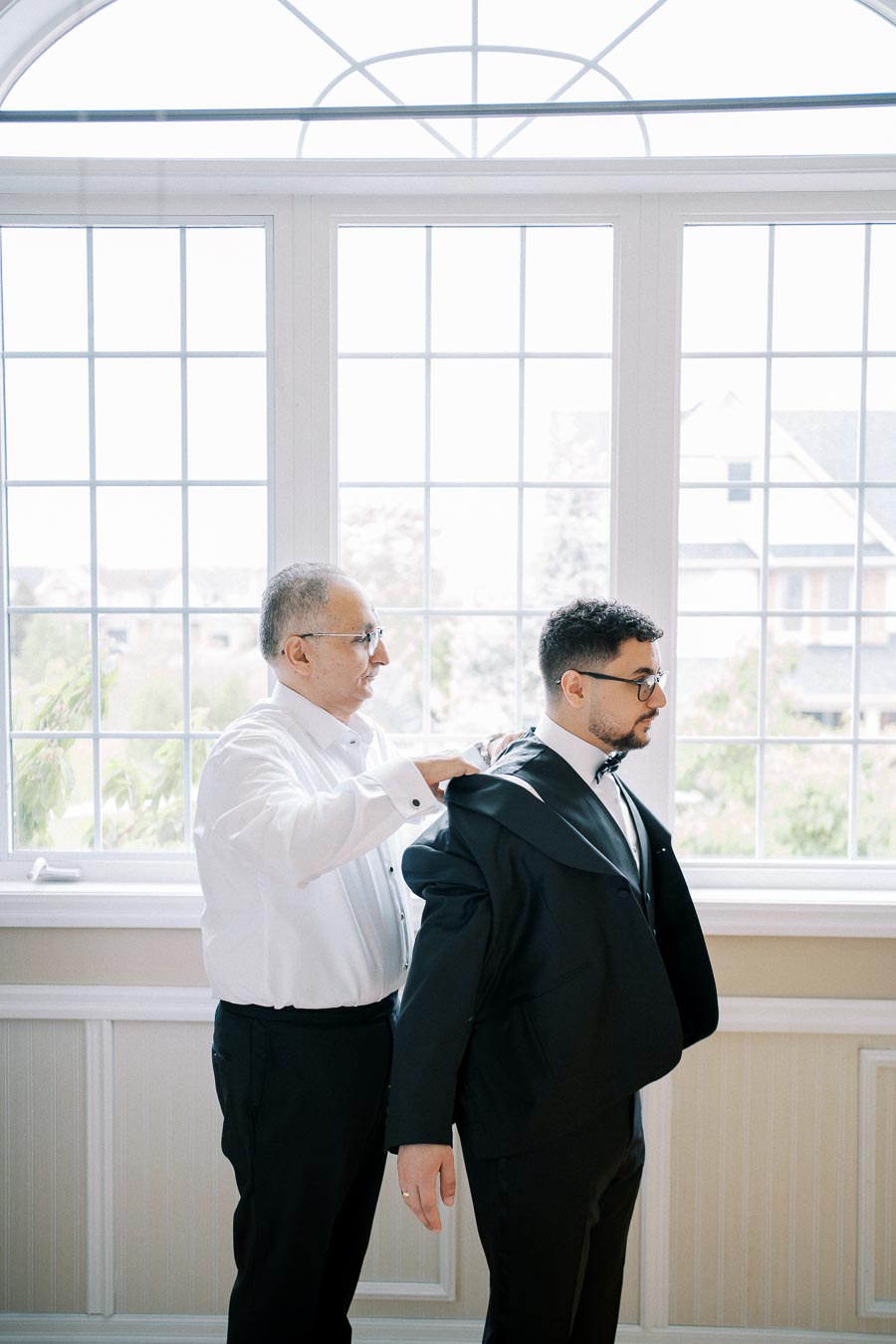 A man helping another man put on a suit jacket in a bright room with large windows and a scenic view outside, focusing on preparation for a formal event or wedding.