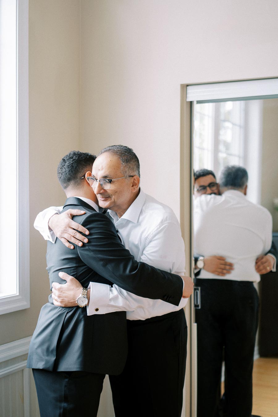 Father and son sharing an emotional hug by a window, reflecting in a nearby mirror, dressed in formal attire.