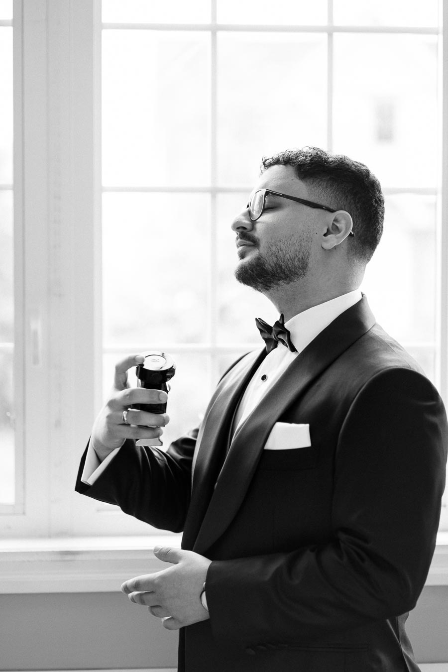 Black and white image of a man in a tuxedo applying cologne, standing by a window with soft natural light.