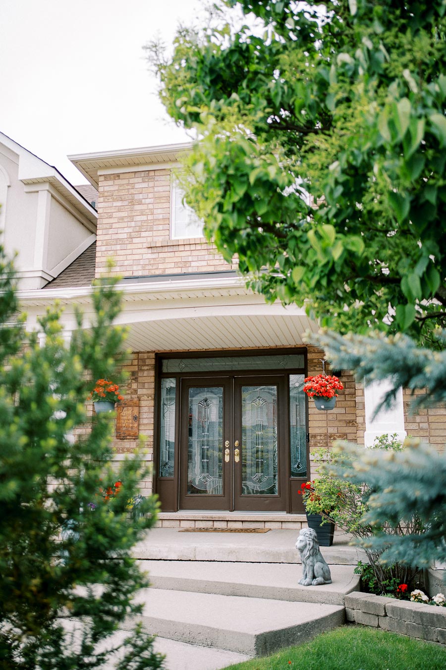 Alt text: Charming brick house entrance with double doors, surrounded by lush greenery and blooming flowers, featuring a decorative lion statue.