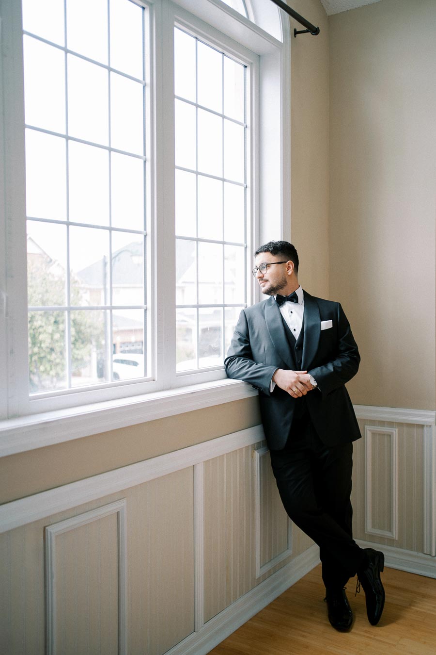 Man in a black tuxedo looking out a large window, daylight illuminating the elegant room interior.