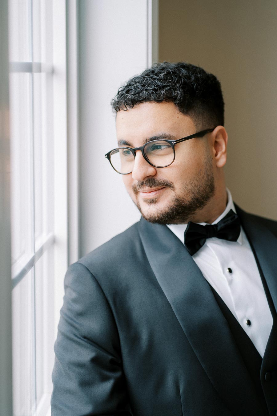 A person in a tuxedo and glasses gazes out a window, exuding confidence and elegance.