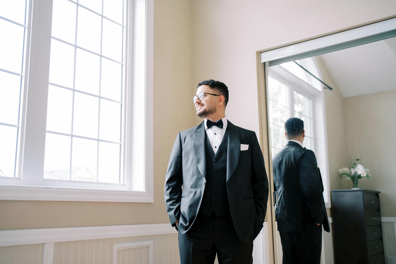 A man in a formal black tuxedo with a bow tie stands confidently in a sunlit room, looking out a large window. His reflection is visible in an adjacent mirror, showcasing the bright, elegant setting and interior decor.
