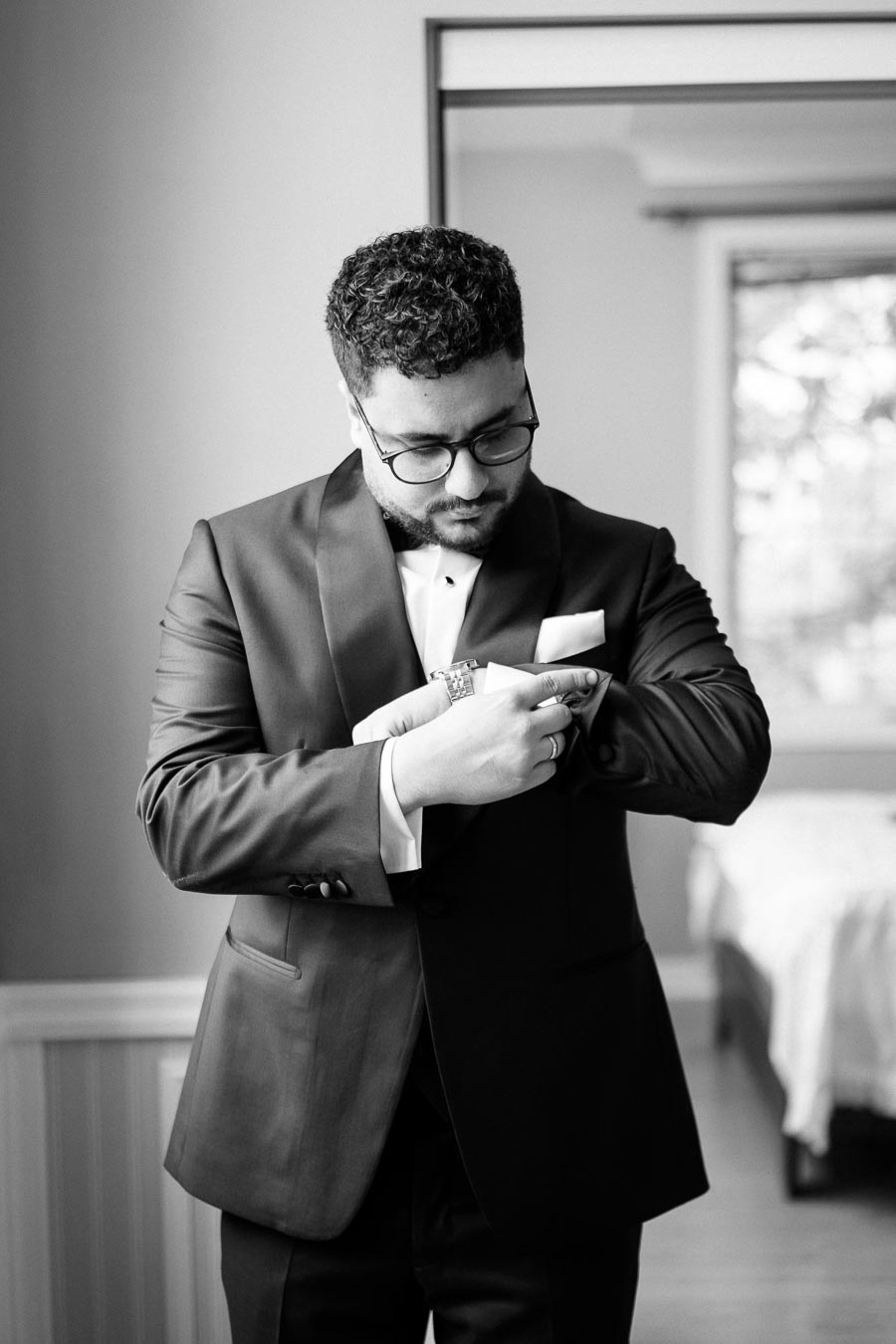 A man dressed in a formal black suit adjusts his cufflinks in a well-lit room. The photograph, captured in black and white, conveys a sense of elegance and preparation, ideal for a sophisticated event or wedding.