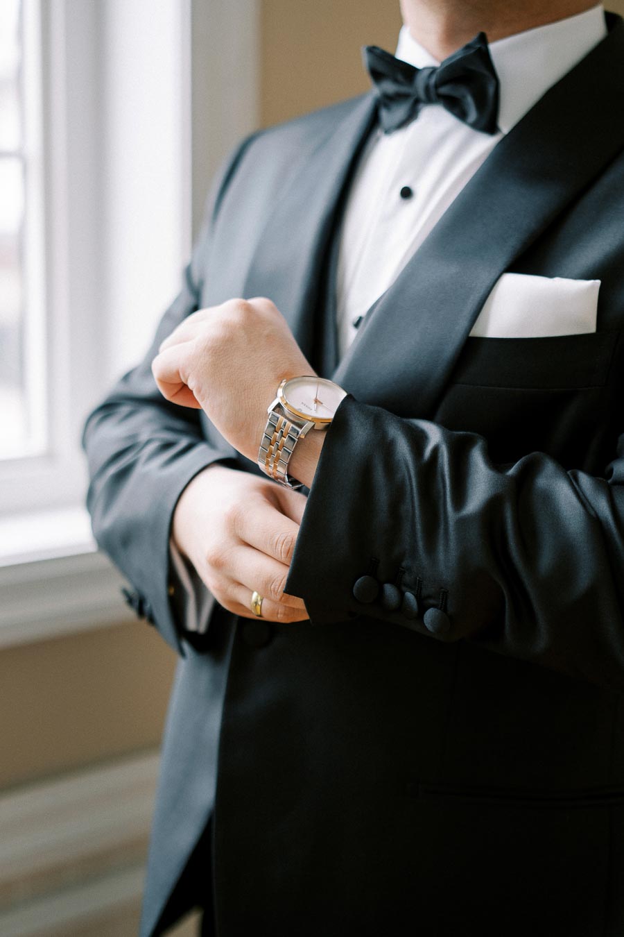 A man in a black tuxedo adjusting his silver wristwatch, standing beside a window, showcasing elegance and style.