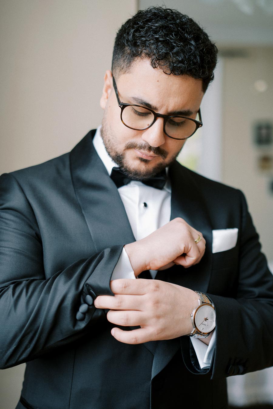 A man in a formal black tuxedo adjusts his cufflinks, showcasing a stylish watch and black-rimmed glasses, preparing for an elegant event.