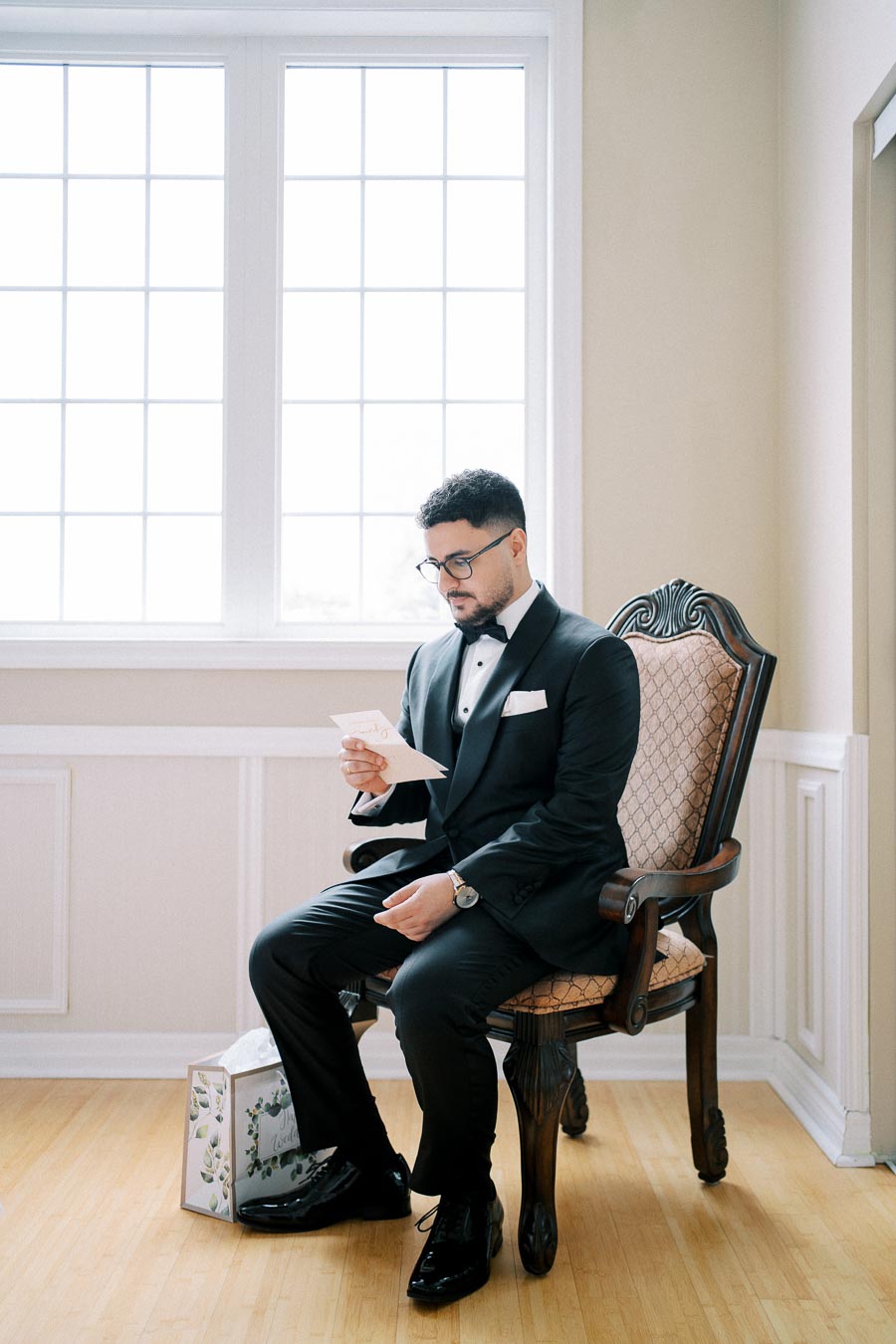 A groom in a black tuxedo sits on an elegant chair reading a letter, with a gift box on the floor beside him, in a well-lit room with large windows.