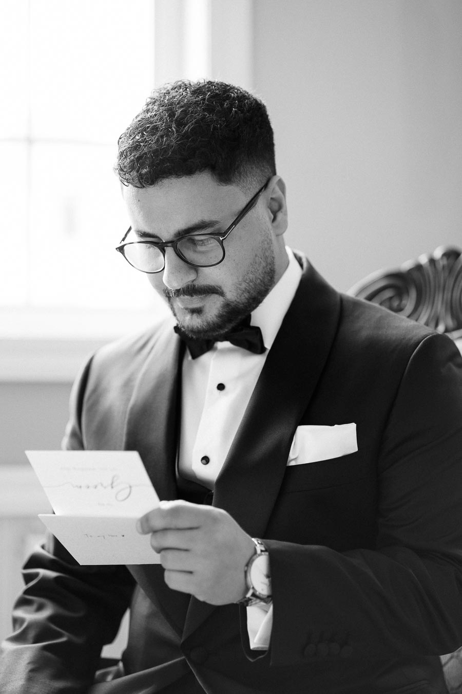 A groom in a tuxedo reading a letter before his wedding day, captured in black and white photography.