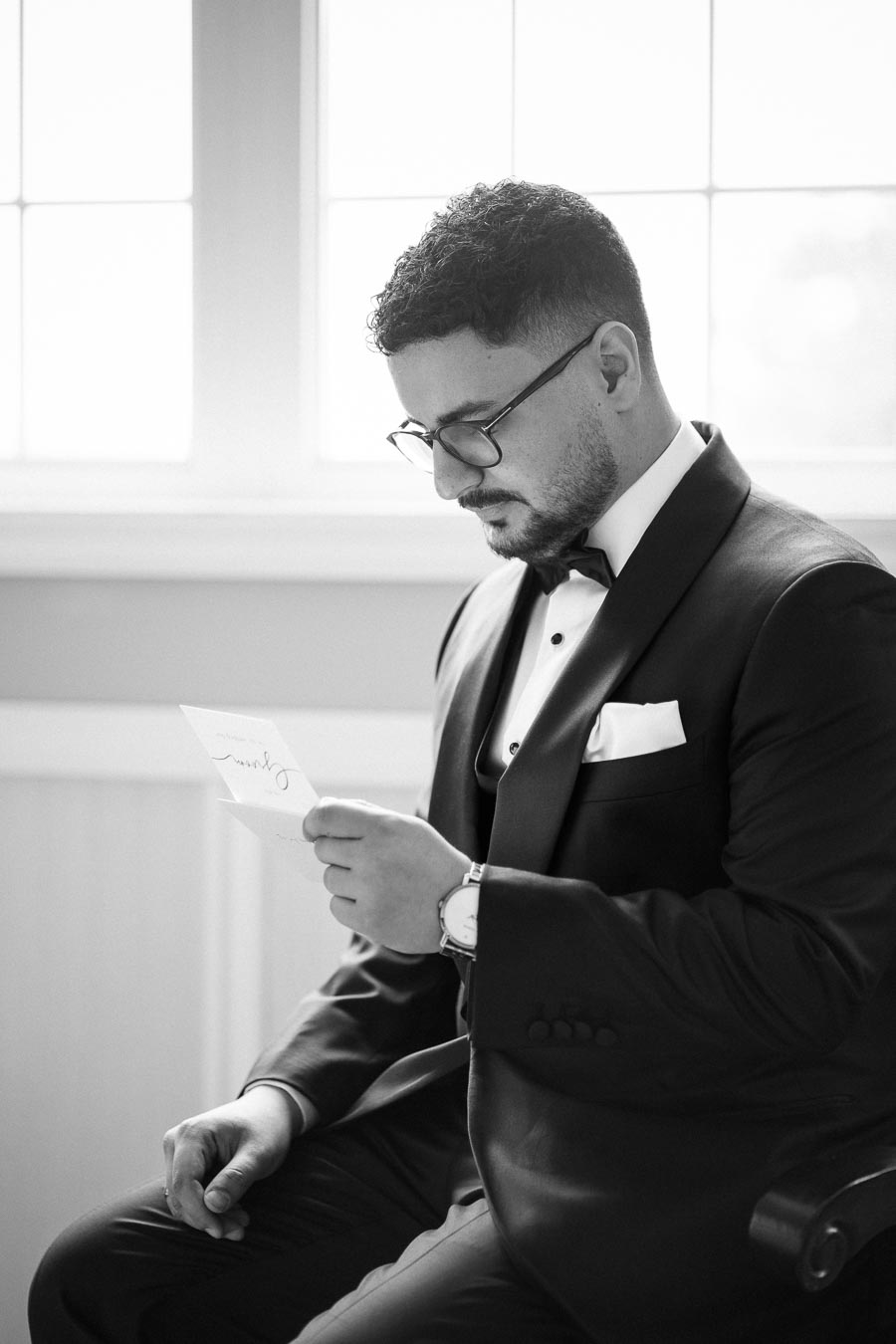 A groom in black-tie attire reads a note, seated in a bright room with soft natural light filtering through a window.