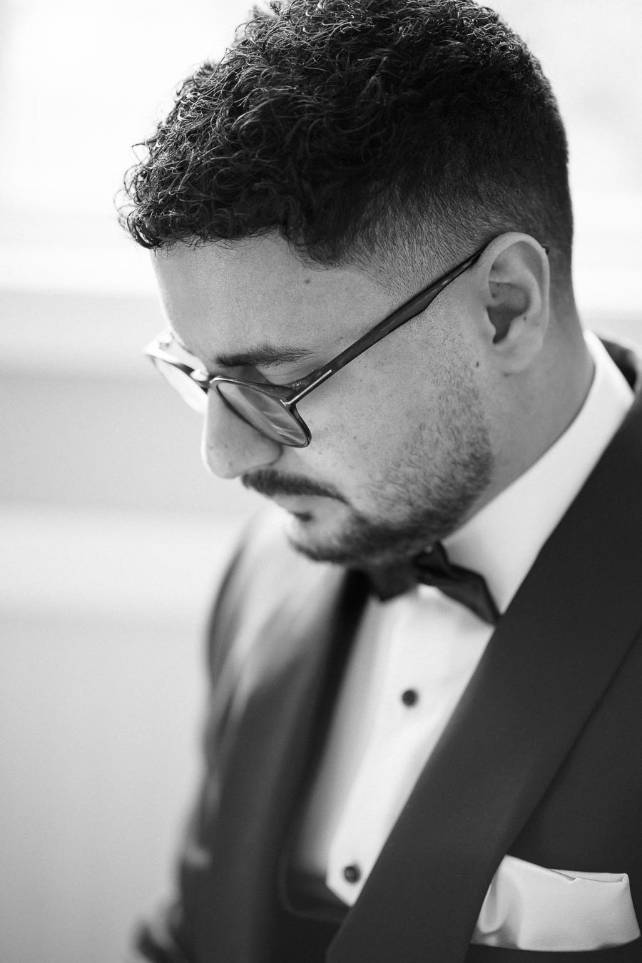 Black and white portrait of a man in glasses wearing a tuxedo, focusing downward with a pensive expression.