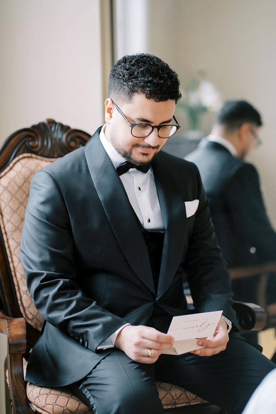 A groom in a black tuxedo sits on a chair, reading a letter.