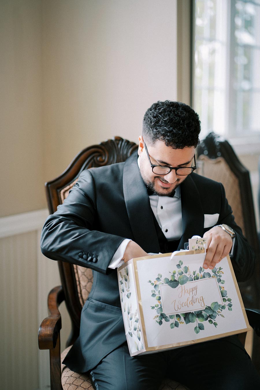 Groom in formal tuxedo opens a Happy Wedding gift bag, seated on an ornate chair by a bright window.