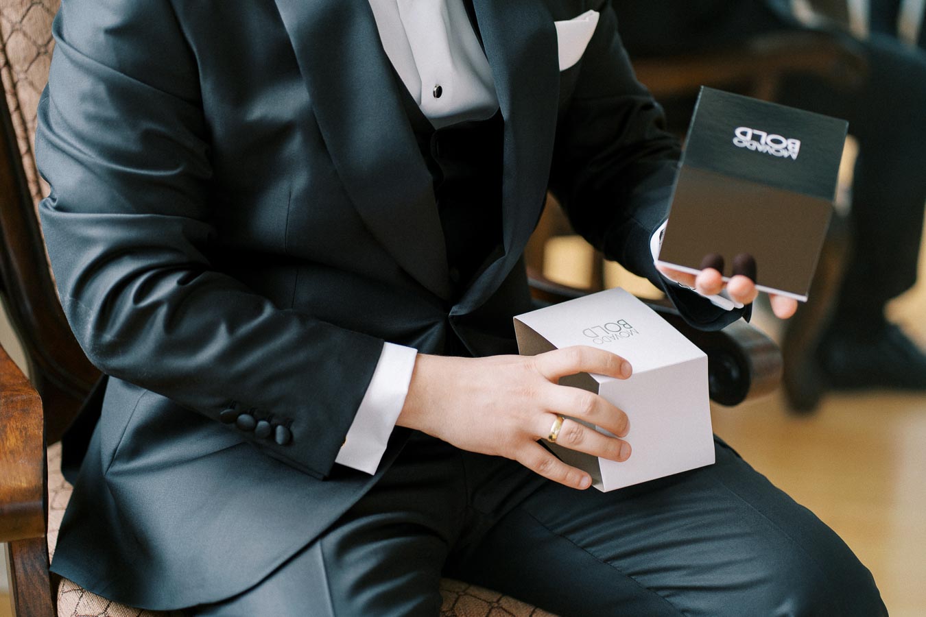 Elegant man in a black suit holding a small gift box, seated on a chair, showcasing luxury packaging, perfect for formal events or special occasions.