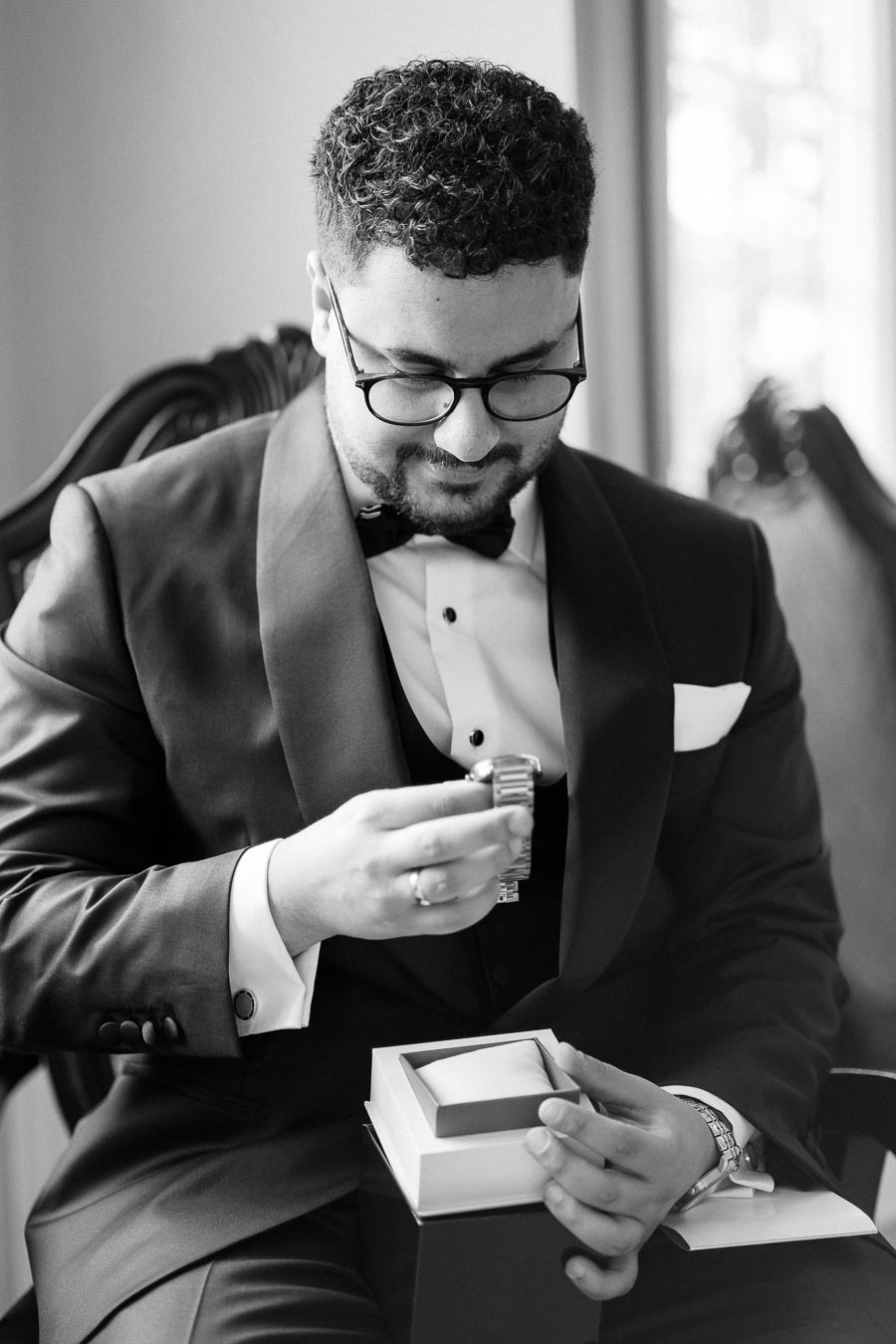 A man dressed in a formal suit, sitting and examining a watch, with an open watch box on his lap. The black and white image captures a thoughtful moment, emphasizing sophistication and elegance.