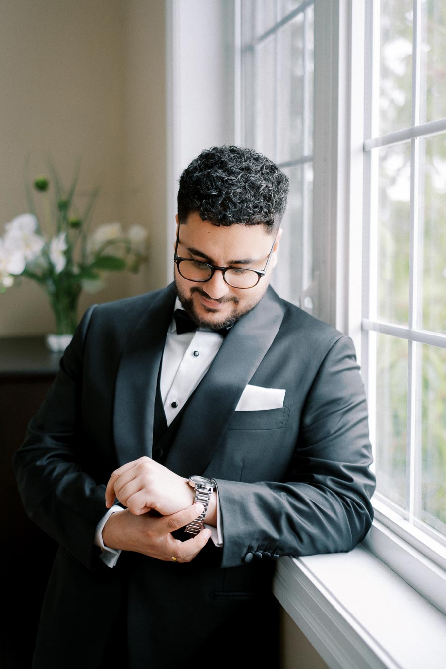 A man in a formal black tuxedo with a bow tie stands by a window, adjusting his wristwatch. He has curly hair, glasses, and a neatly folded pocket square. In the background, a vase with white flowers is visible.