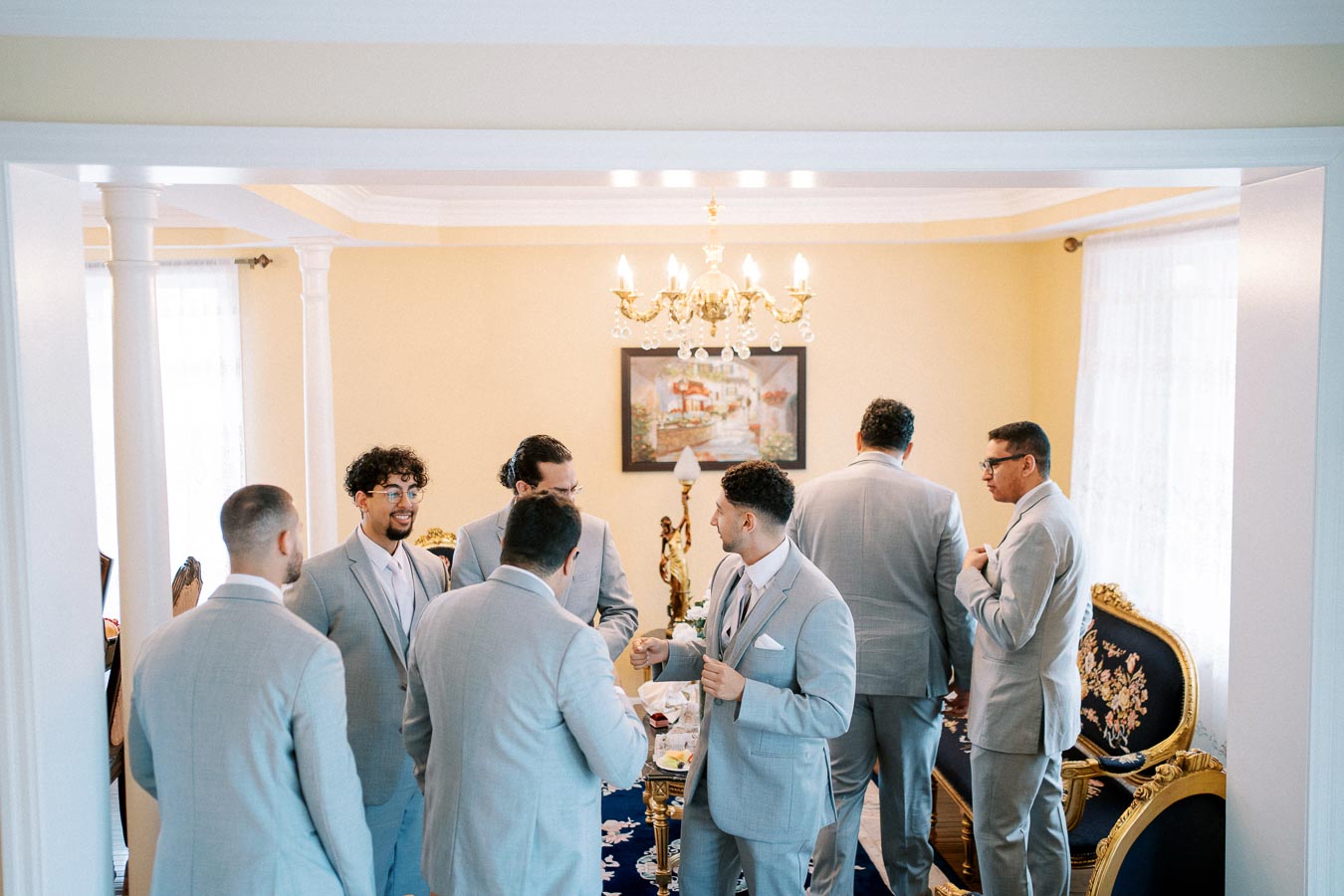 A group of groomsmen in light gray suits gathers in a warmly lit, elegantly decorated room with a chandelier, preparing for a formal event.