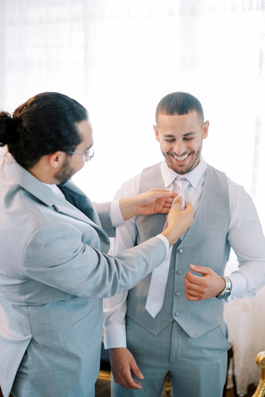 Two men in light gray suits smiling as one helps the other adjust his tie, preparing for a formal event, capturing the joyful moment of camaraderie and friendship.