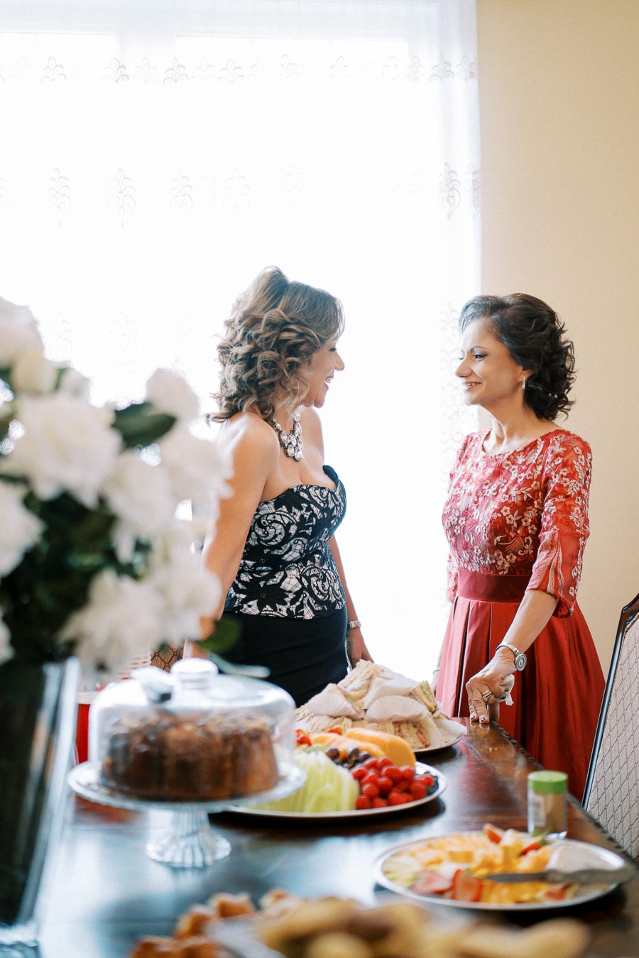 Two women in elegant dresses engaged in conversation at a dining table with an assortment of food including cake and sandwiches, accompanied by floral decorations. The setting suggests a festive or celebratory gathering.