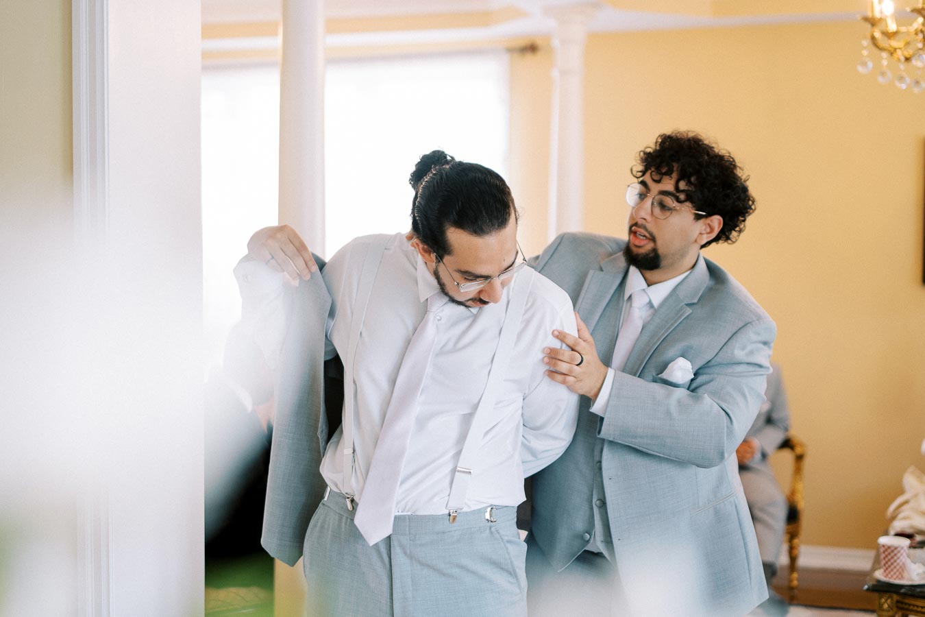 Two men in formal attire, one helping the other put on a light gray suit jacket, preparing for a wedding ceremony indoors.