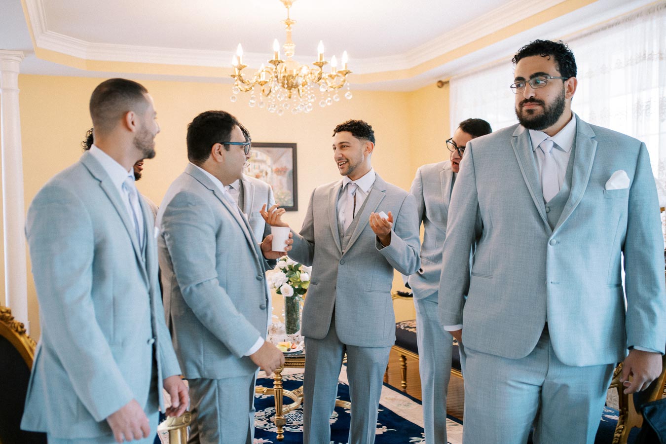 A group of groomsmen in light gray suits chatting and laughing in a well-decorated living room with a chandelier overhead.