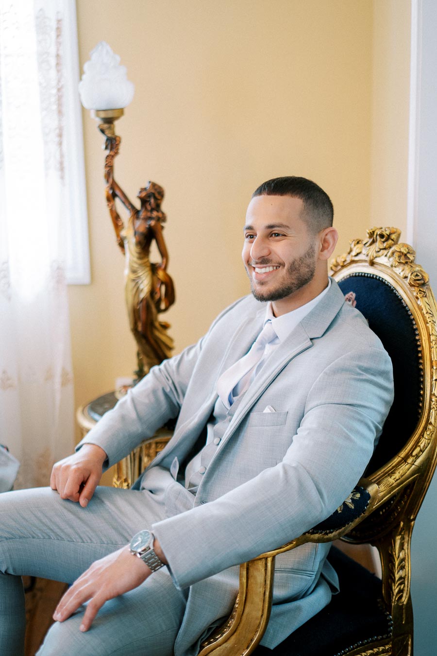 Smiling man in a light gray suit seated on an elegant ornate chair, with soft natural light from a nearby window accentuating the room's classic decor.