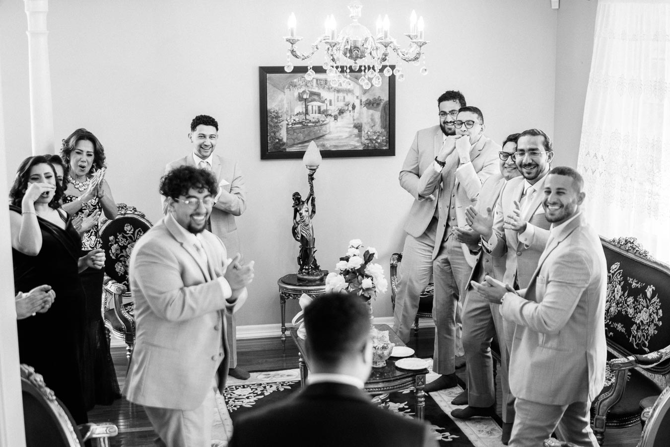 A joyful group of groomsmen in suits, clapping and celebrating indoors around a groom, surrounded by elegant decor and a chandelier.