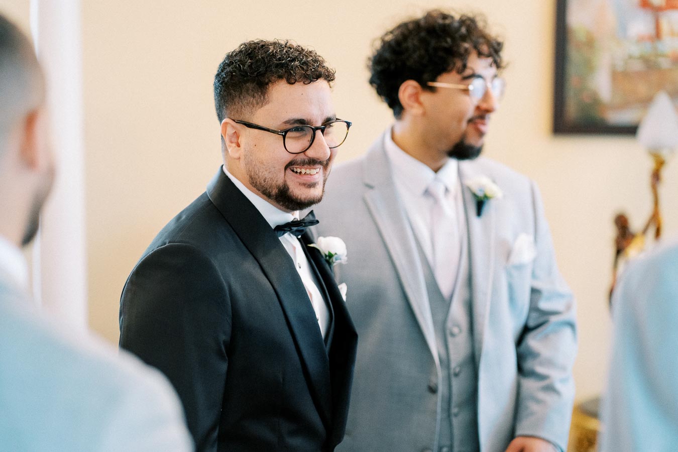 Two men dressed in formal suits, one in a black tuxedo and the other in a light gray suit, smiling and conversing during a wedding event.