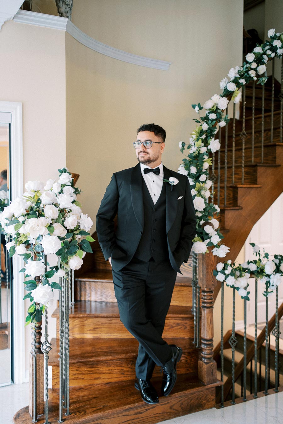 Young man in a black tuxedo with a bow tie standing on a wooden staircase adorned with white floral arrangements, looking confidently to the side in an elegant indoor setting.
