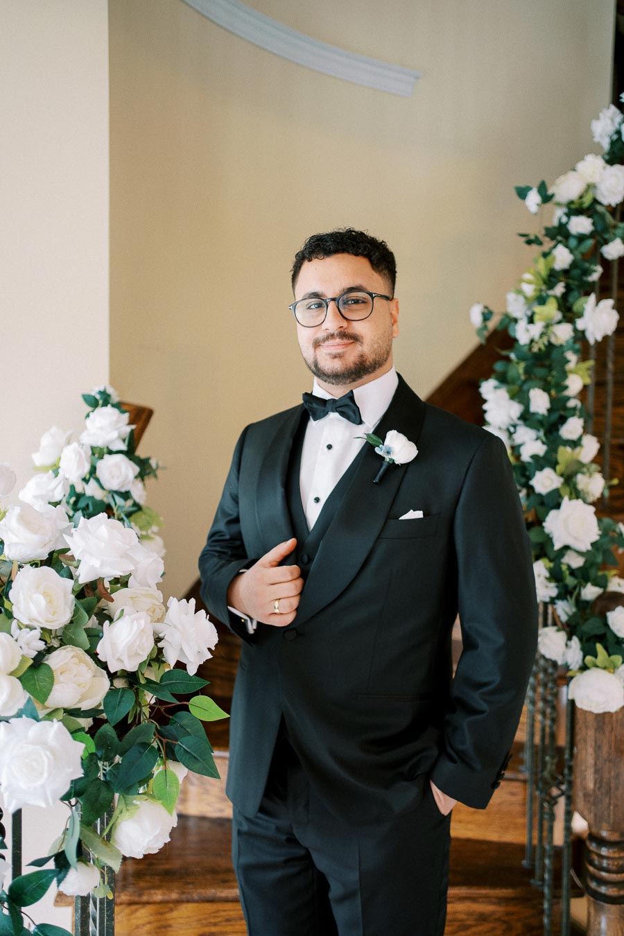 Groom in a black tuxedo with white floral boutonniere standing beside a staircase decorated with white roses.