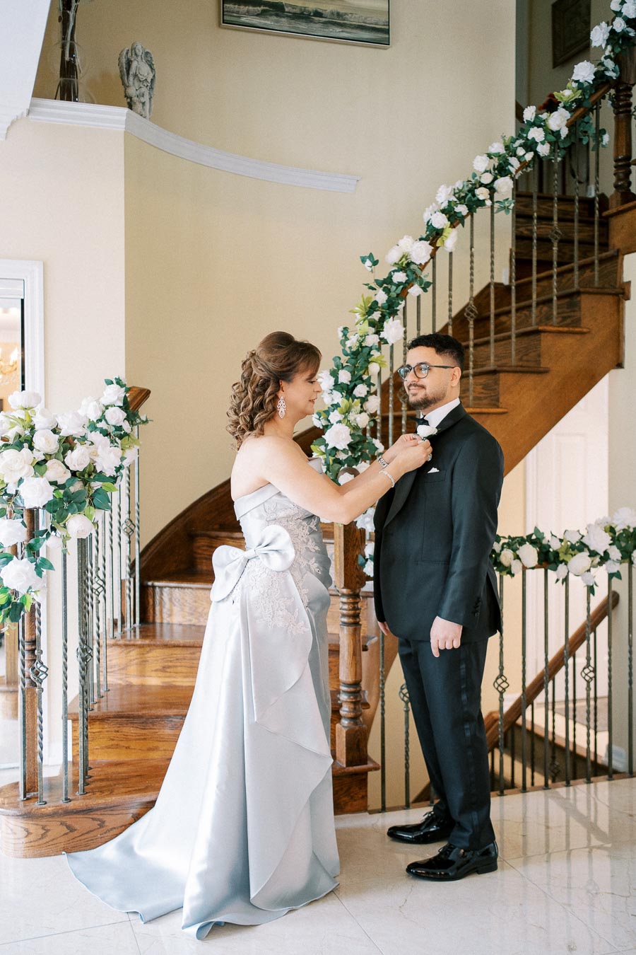 A woman in an elegant silver gown adjusts a boutonniere on a man in a black suit, standing by a beautifully decorated staircase with white floral arrangements.