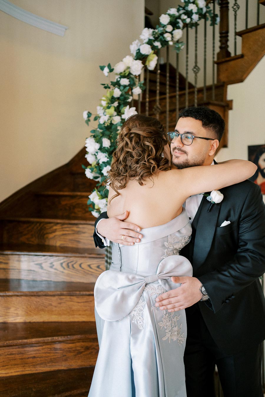 A woman in a light blue dress with a large bow on the back is embracing a man in a black suit and glasses. They are standing on a wooden staircase adorned with white flowers.