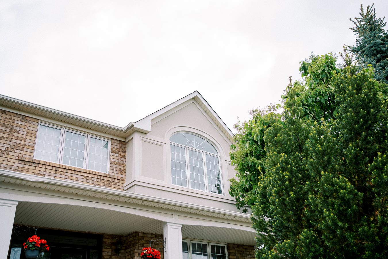 Upper section of a residential house showcasing brick and light-colored siding, large arched windows, and a well-maintained garden with green trees. Ideal for illustrating suburban architecture and home design.