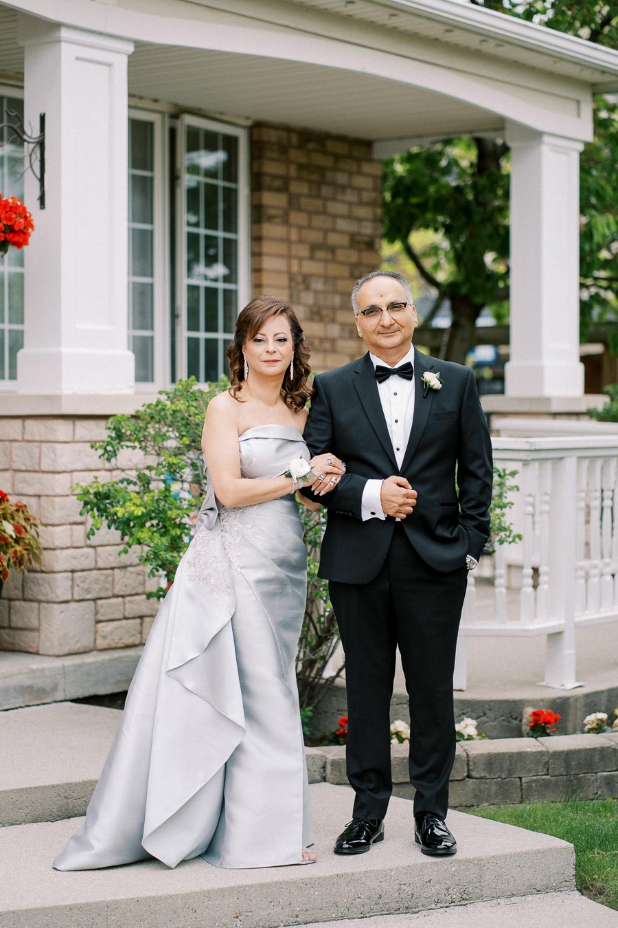 Elderly couple in formal attire posing happily in front of a brick home with a porch, surrounded by greenery and flowers.