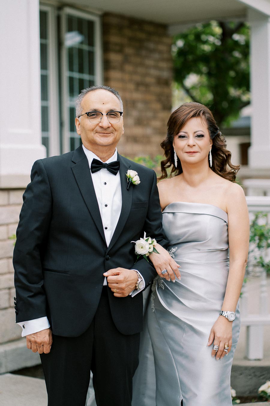 Elegant couple in formal attire, man in a black suit with a bow tie and woman in a silver dress, posing outdoors at a formal event.