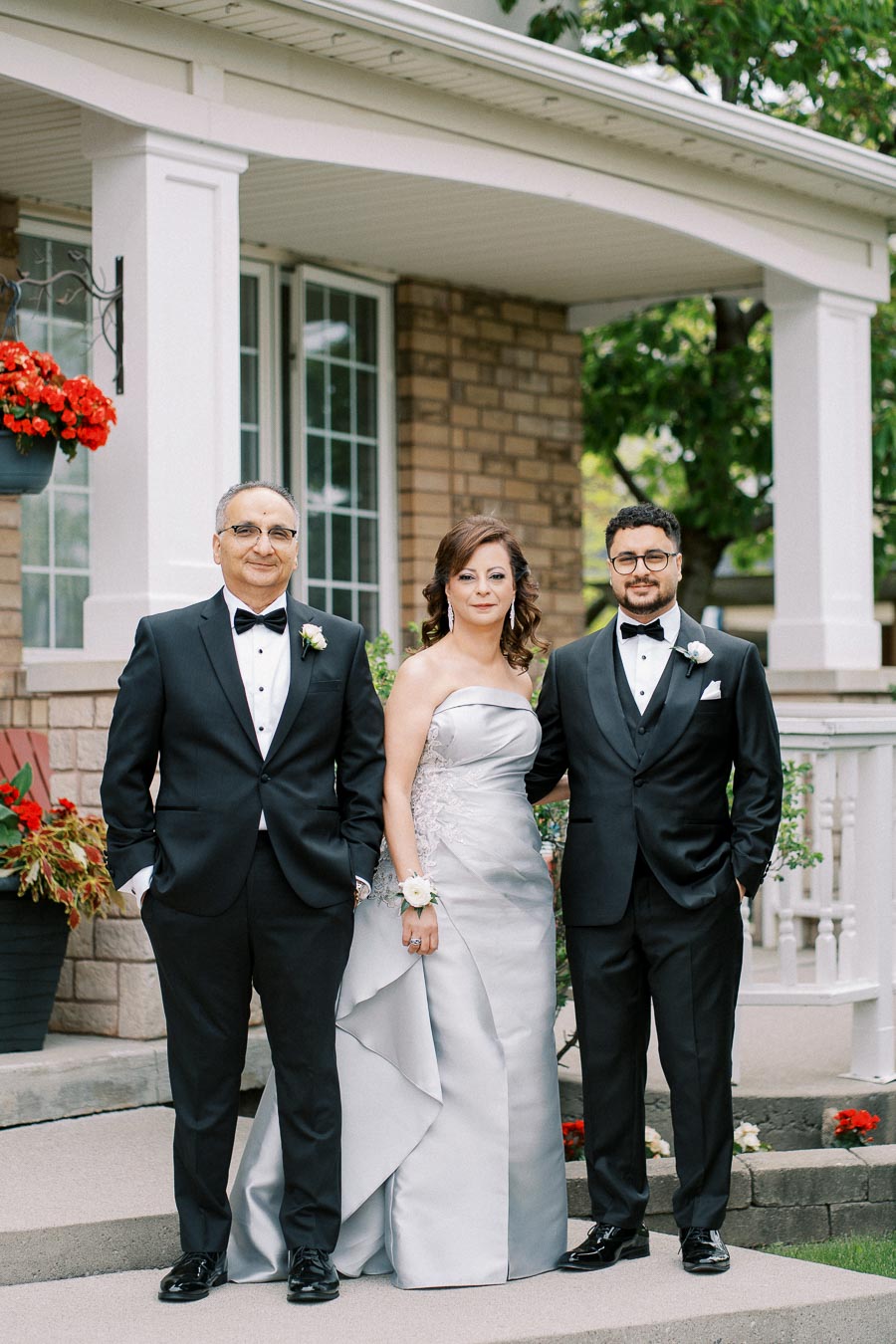 Elegant wedding portrait with three people dressed formally, posing in front of a house with blooming flowers and greenery. The two men in tuxedos and a woman in a silver gown create a sophisticated group photo.