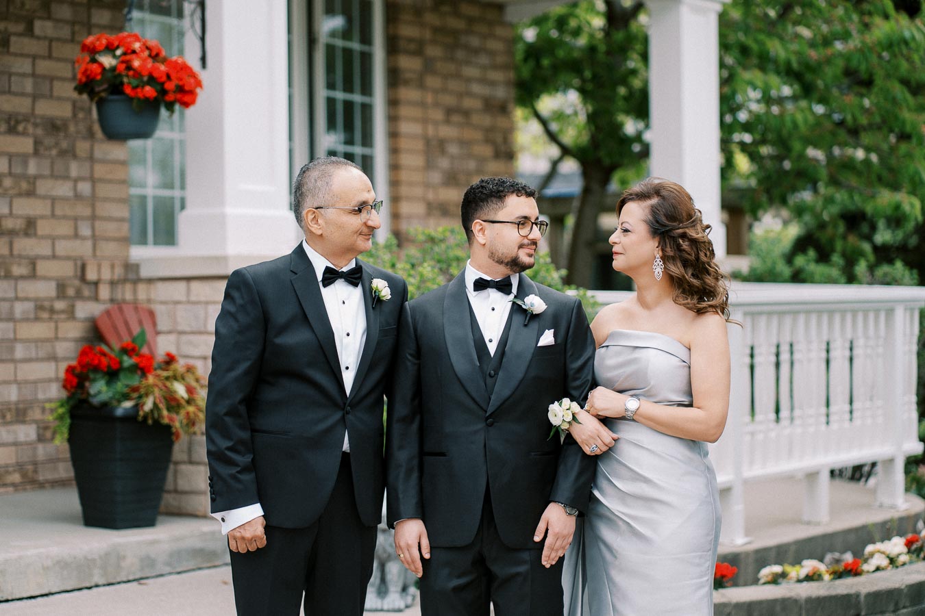 A well-dressed family posing for a photo at an outdoor wedding ceremony, with two men in black tuxedos and a woman in a silver gown, standing in front of a brick house with red flowers.