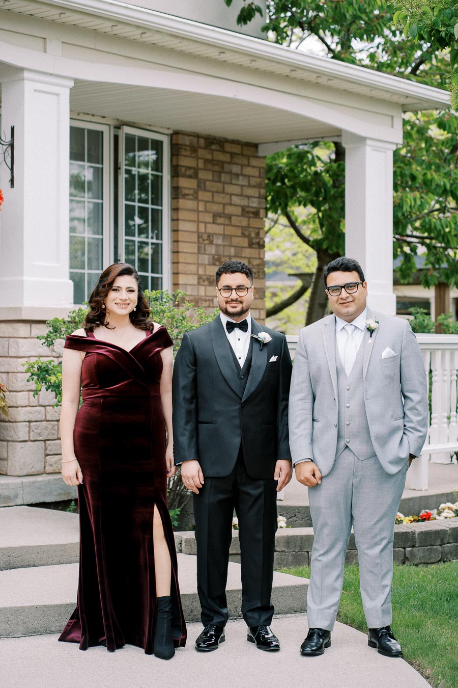 Three well-dressed individuals posing in front of a house, featuring a woman in a burgundy velvet gown with a slit, and two men in formal suits, one in black and the other in light gray. The setting is an outdoor garden with greenery and a brick pathway.
