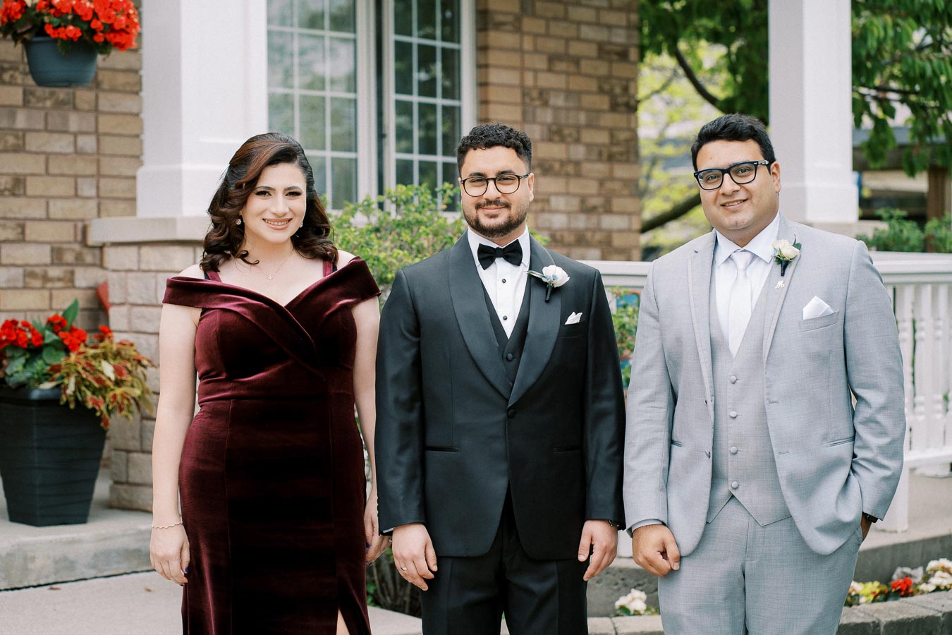 Three people dressed in formal attire standing outside; a woman in a burgundy dress and two men in suits, one in black and the other in gray, with a backdrop of a brick house and flower arrangements.
